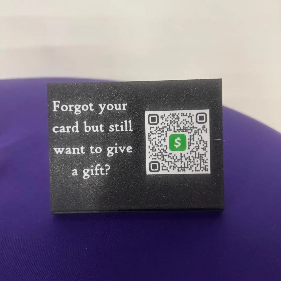 A small black rectangular card with white text reading 'Forgot your card but still want to give a gift?' next to a QR code containing a green square with an S, placed on a purple surface.